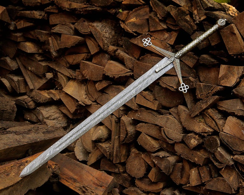 May include: A double-edged sword featuring a distinct wavy Damascus steel pattern on the blade. The silver-toned metal hilt includes a crossguard with four-lobed circular terminals at each end and a textured grip leading to a round pommel.