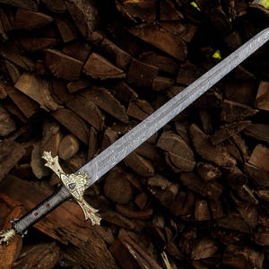 May include: A sword with a long, double-edged blade featuring a distinct wavy Damascus steel pattern. The hilt includes a gold-toned crossguard with ornate detailing, red accents, a black textured grip, and a decorative pommel at the base.