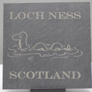 Könnte beinhalten: Ein graues Schiefer-Schild mit den Worten "Loch Ness" und "Schottland" in weißer Schrift. In der Mitte des Schildes befindet sich eine Comic-Abbildung des Seeungeheuers von Loch Ness.
