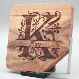 Könnte beinhalten: Eine Holzplakette mit einem dekorativ gravierten Buchstaben K und dem Namen "Kay" darunter. Die Plakette ist braun und hat eine natürliche Holzmaserung. Sie steht auf einem schwarzen Ständer.