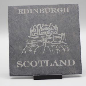 Könnte beinhalten: Ein quadratischer, grauer Schieferuntersetzer mit den weißen Worten "EDINBURGH" und "SCOTLAND". Eine detaillierte Illustration eines Schlosses ist ebenfalls eingraviert. Der Untersetzer wird auf einem schwarzen Ständer präsentiert.