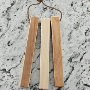 May include: A set of three wooden wind chimes with a leather cord for hanging. The chimes are rectangular, with varying shades of light and medium brown. The center chime is a lighter, natural wood color.