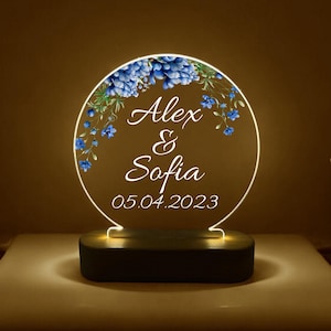 Könnte beinhalten: Ein rundes, weißes LED-Nachtlicht mit einem Holzsockel. Das Licht zeigt ein blaues Blumenmuster und den Text "Alex & Sofia 05.04.2023".