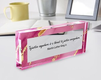 May include: A rectangular, pink acrylic block with gold veining and the quote "Injustice anywhere is a threat to justice everywhere." by Martin Luther King Jr. The block sits on a white surface.