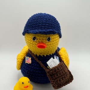 Op de afbeelding: Een gehaakte gele eend met een marineblauwe hoed en uniform, met een kleine Amerikaanse vlagspeld. Het heeft een bruine tas en een kleinere rubberen eend in de buurt. De eend heeft zwarte ogen en een oranje snavel.