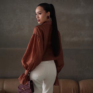 Puede incluir: Una mujer con una blusa color &oacute;xido de mangas voluminosas y pantalones blancos. Sostiene un bolso burdeos con un dise&ntilde;o tejido. La mujer tiene pelo largo y negro y lleva pendientes grandes y redondos.