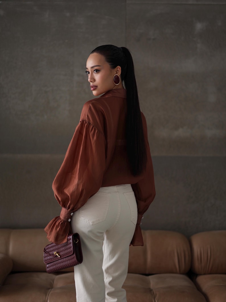 Puede incluir: Una mujer con una blusa color &oacute;xido de mangas voluminosas y pantalones blancos. Sostiene un bolso burdeos con un dise&ntilde;o tejido. La mujer tiene pelo largo y negro y lleva pendientes grandes y redondos.