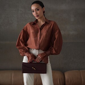 Puede incluir: Una mujer con una camisa abotonada color cobre con mangas voluminosas y pantalones blancos. Sostiene un bolso de mano burdeos oscuro con un cierre dorado. El fondo es una pared gris neutra y un sof&aacute; de cuero marr&oacute;n.