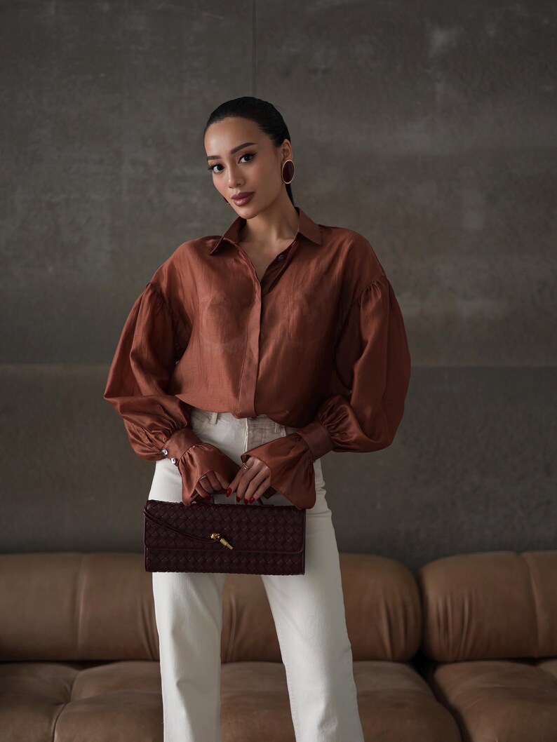 Puede incluir: Una mujer con una camisa abotonada color cobre con mangas voluminosas y pantalones blancos. Sostiene un bolso de mano burdeos oscuro con un cierre dorado. El fondo es una pared gris neutra y un sof&aacute; de cuero marr&oacute;n.