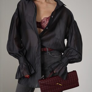 Puede incluir: Una camisa abotonada gris oscuro y transparente con mangas con volantes. Un sujetador burdeos es visible debajo de la camisa abierta. El conjunto se combina con unos vaqueros grises y un clutch burdeos.