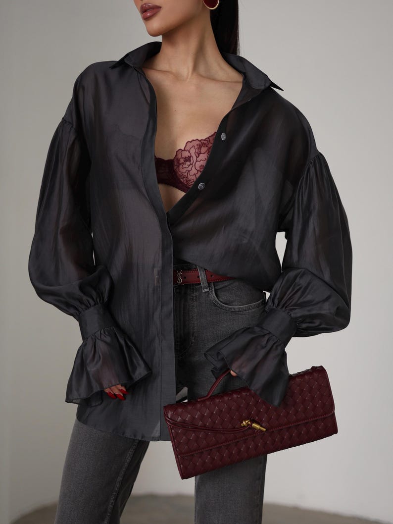 Puede incluir: Una camisa abotonada gris oscuro y transparente con mangas con volantes. Un sujetador burdeos es visible debajo de la camisa abierta. El conjunto se combina con unos vaqueros grises y un clutch burdeos.