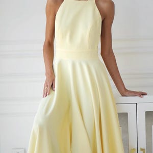 Pu&ograve; includere: Abito giallo pallido con collo all'americana e gonna svasata. L'abito ha un corpetto aderente e un'ampia cintura. Il tessuto sembra essere un materiale liscio e fluido. L'abito &egrave; lungo fino al ginocchio ed &egrave; adatto alle occasioni formali.