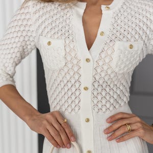 May include: White knit cardigan with a diamond pattern and gold buttons. The cardigan has two small pockets and a V-neck. The outfit includes a matching white dress. The style is elegant and classic.