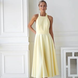 May include: A pale yellow halter dress with a flared skirt. The dress has a fitted bodice and a midi length. The model is wearing white heels. The background is a white wall with architectural details.