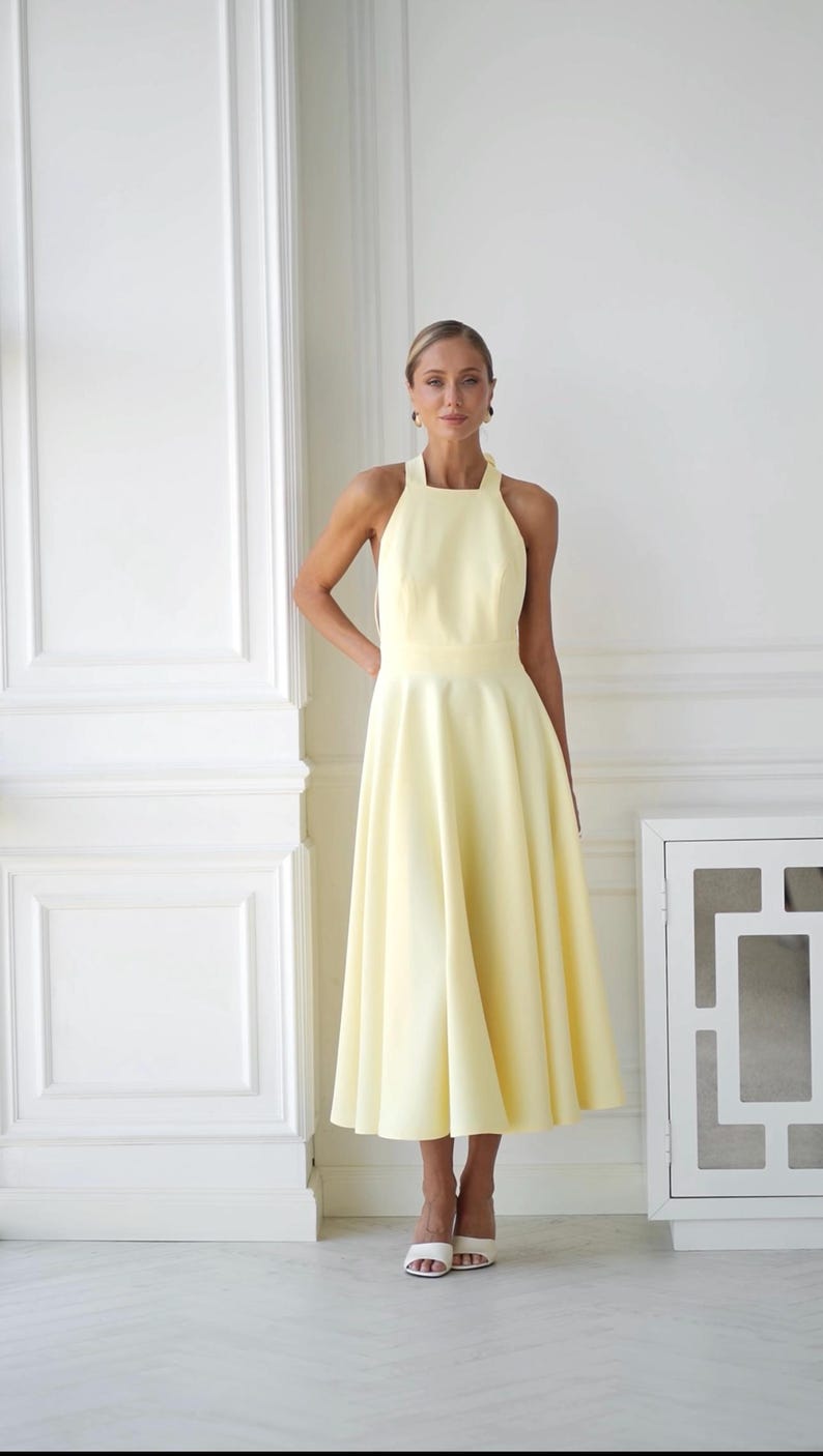 May include: A pale yellow halter dress with a flared skirt. The dress has a fitted bodice and a midi length. The model is wearing white heels. The background is a white wall with architectural details.