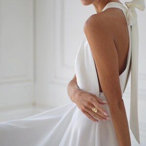 Pu&ograve; includere: Abito bianco con collo all'americana, gonna fluida e dettaglio a fiocco sulla schiena. L'abito presenta una schiena scoperta ed &egrave; indossato con orecchini e un anello dorati. Lo stile generale &egrave; elegante e adatto a eventi formali.