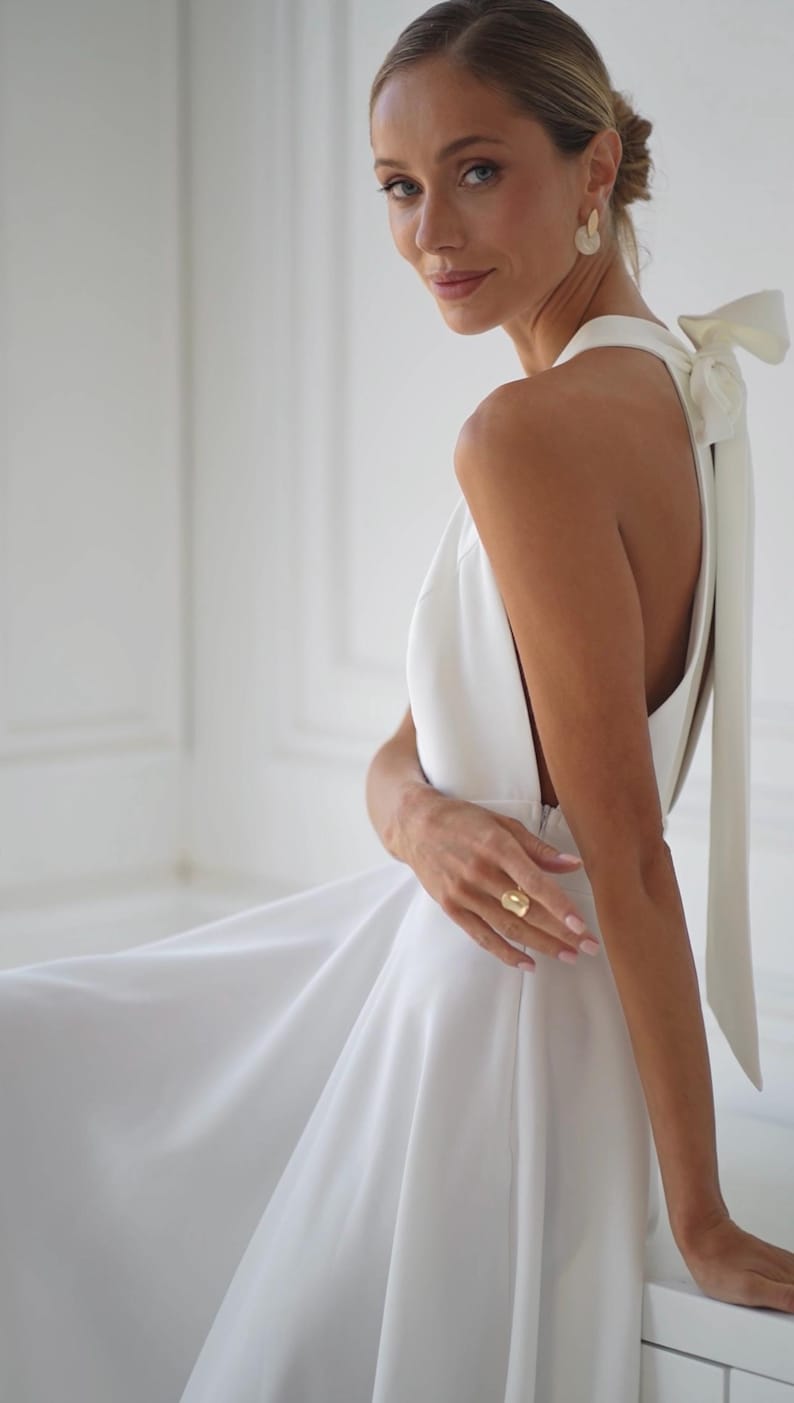 Pu&ograve; includere: Abito bianco con collo all'americana, gonna fluida e dettaglio a fiocco sulla schiena. L'abito presenta una schiena scoperta ed &egrave; indossato con orecchini e un anello dorati. Lo stile generale &egrave; elegante e adatto a eventi formali.