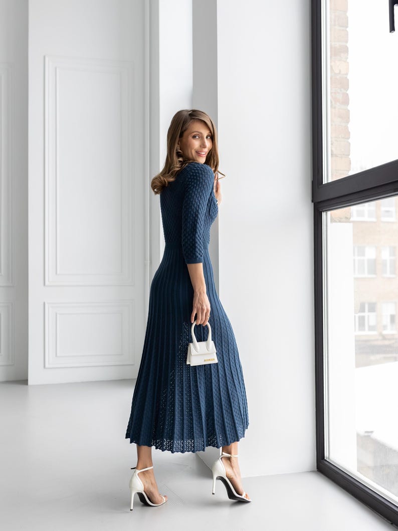 May include: A navy blue, textured knit dress with a pleated skirt and three-quarter sleeves. The dress is paired with white high-heeled sandals and a small white handbag. The outfit is set against a bright, modern interior with a large window.