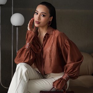 Puede incluir: Una blusa de seda marr&oacute;n con mangas largas y abullonadas y pu&ntilde;os abotonados. La blusa se combina con pantalones color crema y un clutch de cuero trenzado burdeos. Los pendientes de aro dorados completan el look. La est&eacute;tica general es elegante y sofisticada.