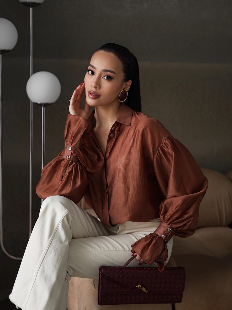 Puede incluir: Una blusa de seda marr&oacute;n con mangas largas y abullonadas y pu&ntilde;os abotonados. La blusa se combina con pantalones color crema y un clutch de cuero trenzado burdeos. Los pendientes de aro dorados completan el look. La est&eacute;tica general es elegante y sofisticada.