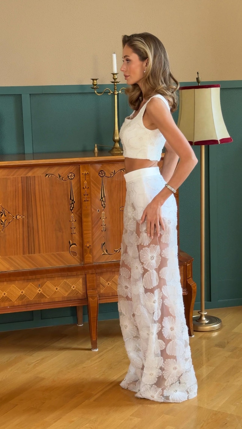 May include: White two-piece outfit featuring a crop top and a long skirt adorned with floral appliqu&eacute;s. The skirt has a sheer overlay. The outfit is suitable for special occasions. The background includes a wooden cabinet and a green wall.
