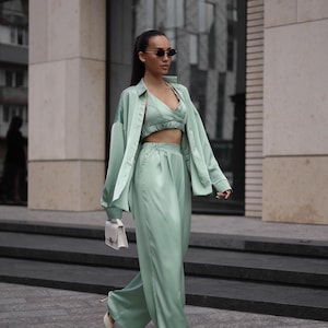 May include: A mint green satin three-piece outfit consisting of a crop top, high-waisted pants, and a button-down shirt. The outfit is paired with a small white handbag and light-colored heels. The model is wearing sunglasses.