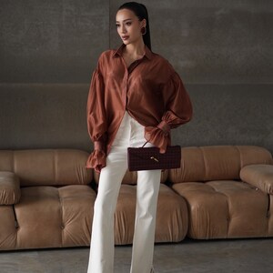 Puede incluir: Una mujer con una blusa de seda color &oacute;xido con mangas voluminosas y vaqueros blancos acampanados. Sostiene un clutch burdeos tejido. El conjunto se combina con tacones burdeos y pendientes de aro dorados. El fondo presenta un sof&aacute; de cuero marr&oacute;n.