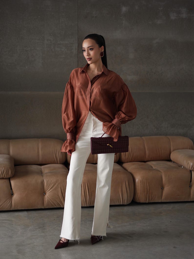 Puede incluir: Una mujer con una blusa de seda color &oacute;xido con mangas voluminosas y vaqueros blancos acampanados. Sostiene un clutch burdeos tejido. El conjunto se combina con tacones burdeos y pendientes de aro dorados. El fondo presenta un sof&aacute; de cuero marr&oacute;n.