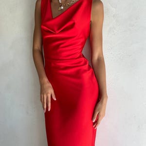 May include: A sleeveless, floor-length red satin dress featuring a draped cowl neckline. The fabric has a smooth, reflective finish. A gold-toned chain necklace with a large white pearl pendant is worn around the neck, resting against the bodice.