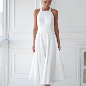 Pu&ograve; includere: Abito midi bianco con scollo all'americana e gonna svasata. L'abito presenta un corpetto aderente e una gonna fluida che arriva a met&agrave; polpaccio. La modella indossa sandali bianchi con tacco. Lo sfondo &egrave; una stanza bianca con una finestra.