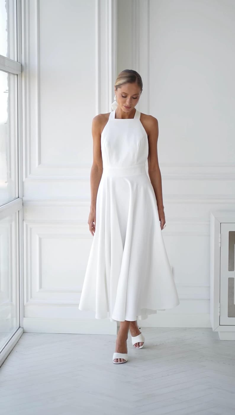 Pu&ograve; includere: Abito midi bianco con scollo all'americana e gonna svasata. L'abito presenta un corpetto aderente e una gonna fluida che arriva a met&agrave; polpaccio. La modella indossa sandali bianchi con tacco. Lo sfondo &egrave; una stanza bianca con una finestra.