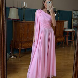 May include: A floor-length, light pink, one-shoulder dress featuring a draped bodice and a long, flowing cape-like sleeve attached at the shoulder. The skirt is full and pleated, falling to the floor in soft, layered fabric folds.
