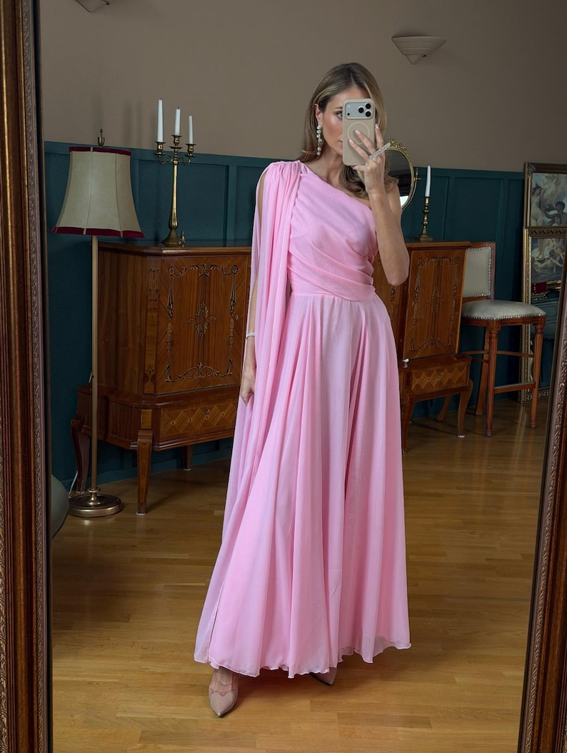 May include: A floor-length, light pink, one-shoulder dress featuring a draped bodice and a long, flowing cape-like sleeve attached at the shoulder. The skirt is full and pleated, falling to the floor in soft, layered fabric folds.