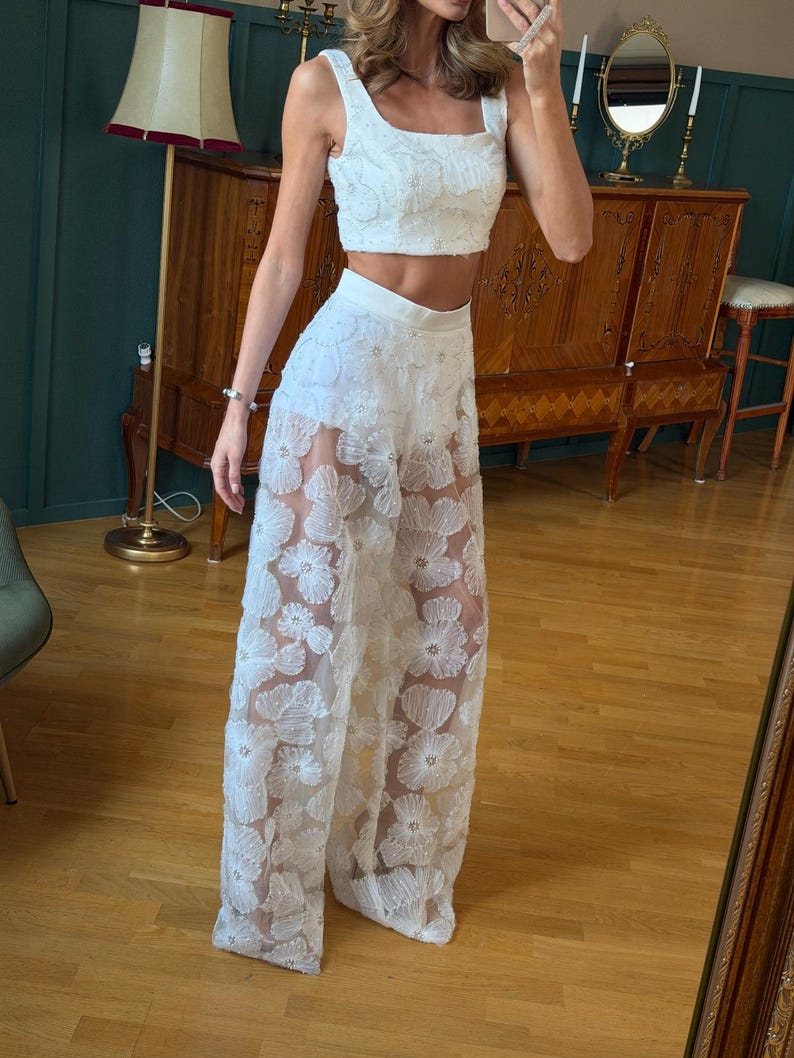 May include: A white two-piece outfit featuring a crop top and wide-leg pants. The top has a square neckline and the pants are adorned with floral embroidery. The outfit is made of a sheer fabric, creating a layered effect.