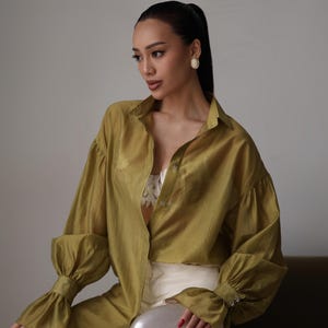 Puede incluir: Una camisa verde oliva con mangas largas y abullonadas, con pu&ntilde;os abotonados. Se combina con pantalones blancos y un clutch plateado. La persona lleva pendientes blancos y el pelo recogido en una cola de caballo.