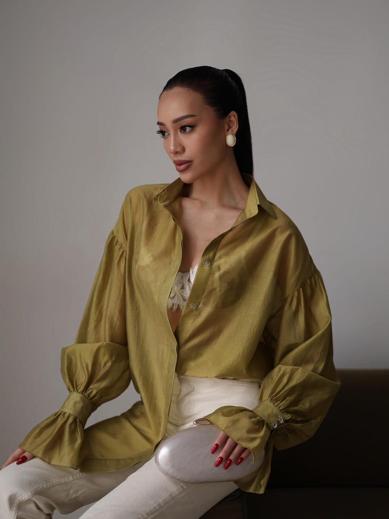 Puede incluir: Una camisa verde oliva con mangas largas y abullonadas, con pu&ntilde;os abotonados. Se combina con pantalones blancos y un clutch plateado. La persona lleva pendientes blancos y el pelo recogido en una cola de caballo.