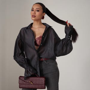 Puede incluir: Una camisa abotonada transparente gris oscuro con mangas largas y un sujetador burdeos. La modelo lleva vaqueros grises y sostiene un clutch burdeos tejido con un cierre dorado. Pendientes burdeos y un cintur&oacute;n burdeos completan el look.