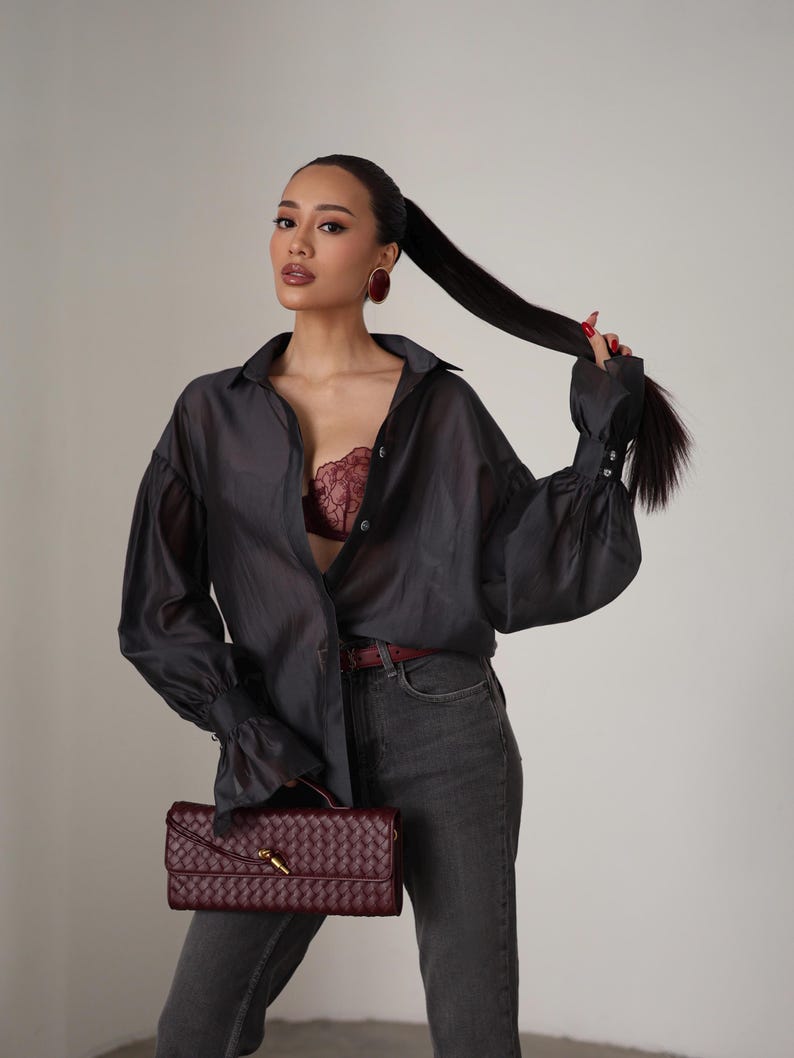 Puede incluir: Una camisa abotonada transparente gris oscuro con mangas largas y un sujetador burdeos. La modelo lleva vaqueros grises y sostiene un clutch burdeos tejido con un cierre dorado. Pendientes burdeos y un cintur&oacute;n burdeos completan el look.