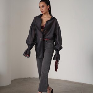 Puede incluir: Una camisa oscura gris transparente con botones y mangas largas y voluminosas, combinada con vaqueros grises y tacones negros. Un cintur&oacute;n y un bolso burdeos a&ntilde;aden un toque de color al conjunto. El conjunto se completa con pendientes burdeos.