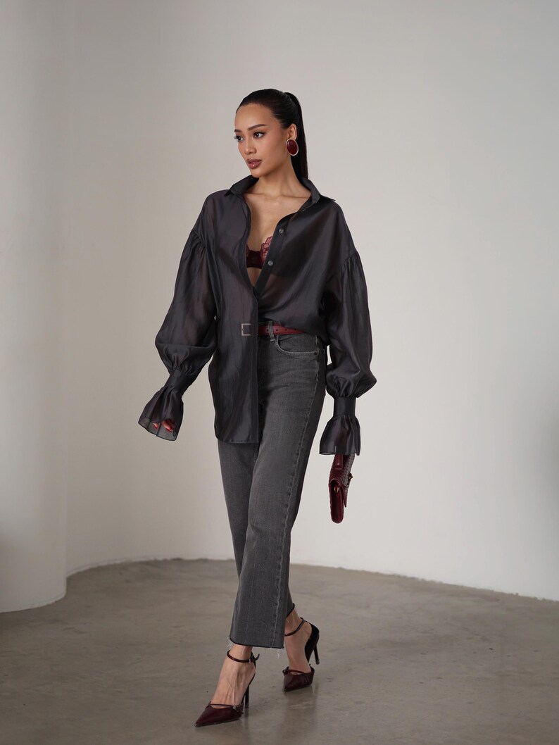 Puede incluir: Una camisa oscura gris transparente con botones y mangas largas y voluminosas, combinada con vaqueros grises y tacones negros. Un cintur&oacute;n y un bolso burdeos a&ntilde;aden un toque de color al conjunto. El conjunto se completa con pendientes burdeos.