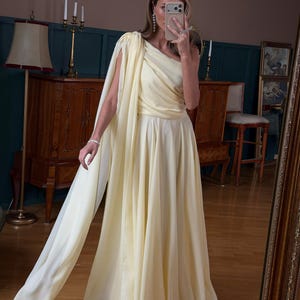 May include: A pale yellow, one-shoulder gown with a flowing skirt and attached cape. The dress features a draped bodice and a floor-length skirt. The fabric appears to be lightweight and flowing, suitable for formal events.