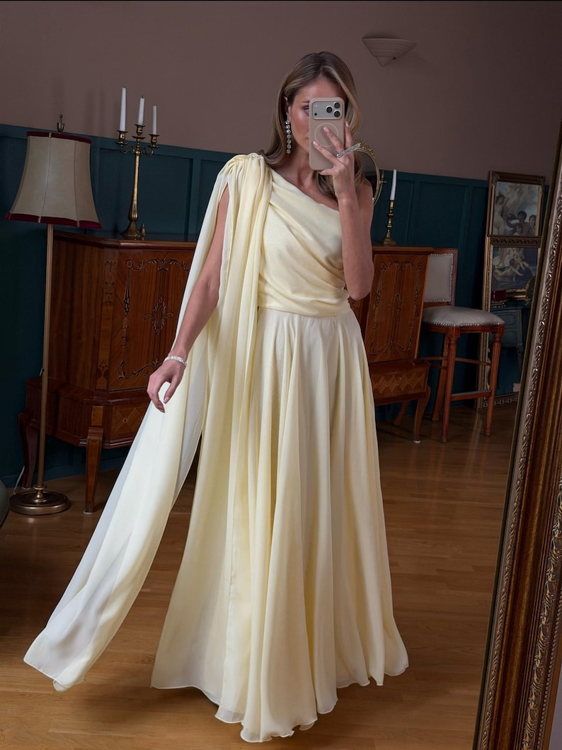May include: A pale yellow, one-shoulder gown with a flowing skirt and attached cape. The dress features a draped bodice and a floor-length skirt. The fabric appears to be lightweight and flowing, suitable for formal events.