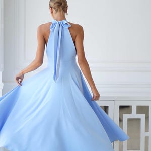 Pu&ograve; includere: Un abito azzurro con collo all'americana e gonna fluida. L'abito presenta un dettaglio a fiocco sulla schiena. La modella indossa tacchi bianchi. L'abito &egrave; in tinta unita e sembra essere realizzato in un tessuto leggero.