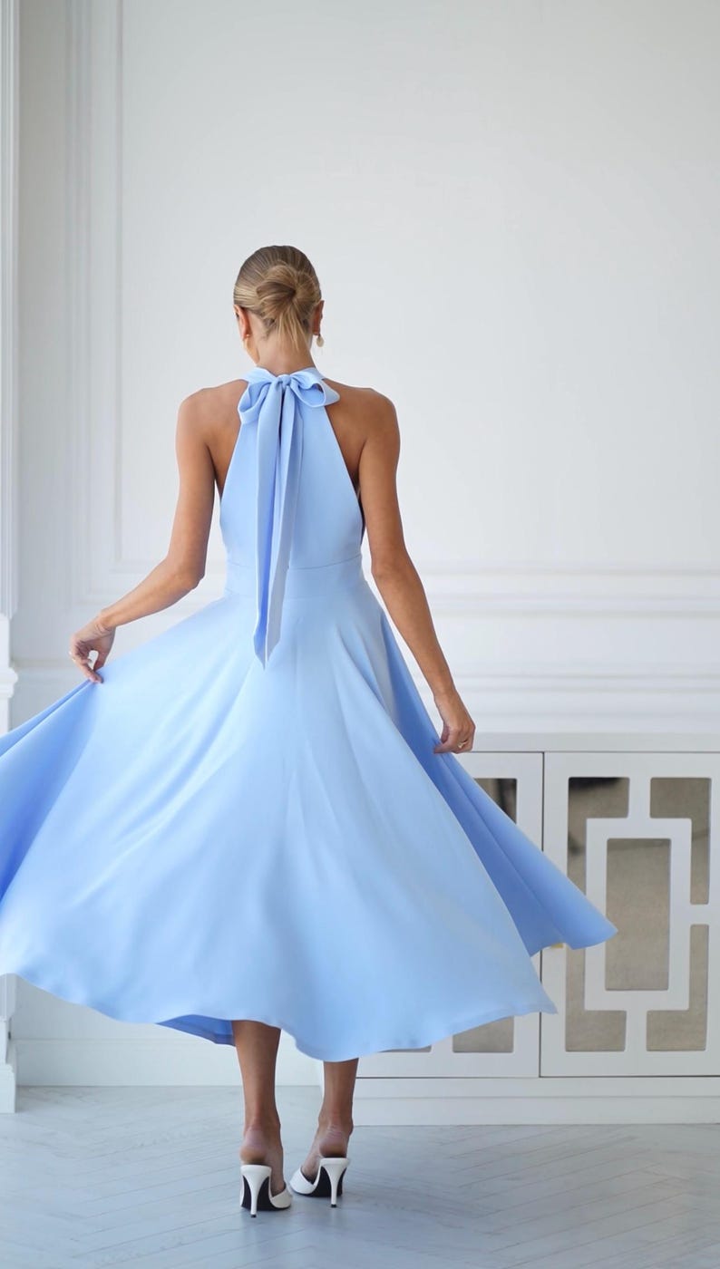 Pu&ograve; includere: Un abito azzurro con collo all'americana e gonna fluida. L'abito presenta un dettaglio a fiocco sulla schiena. La modella indossa tacchi bianchi. L'abito &egrave; in tinta unita e sembra essere realizzato in un tessuto leggero.