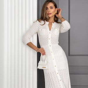May include: A white, open-weave knit dress with a button-down front and a small, white handbag. The dress features a V-neckline, three-quarter sleeves, and gold-colored buttons. The handbag has a top handle and a gold-colored logo.