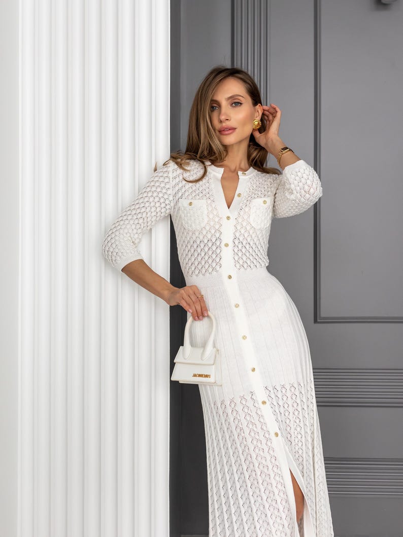 May include: A white, open-weave knit dress with a button-down front and a small, white handbag. The dress features a V-neckline, three-quarter sleeves, and gold-colored buttons. The handbag has a top handle and a gold-colored logo.