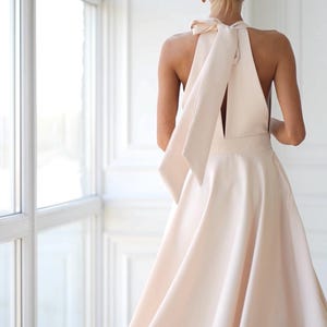 Pu&ograve; includere: Un abito a collo alto color pesca chiaro con gonna fluida e dettaglio a fiocco sulla schiena. L'abito ha una vita aderente e una gonna svasata, creando una silhouette elegante. L'abito &egrave; realizzato in un tessuto liscio e morbido.