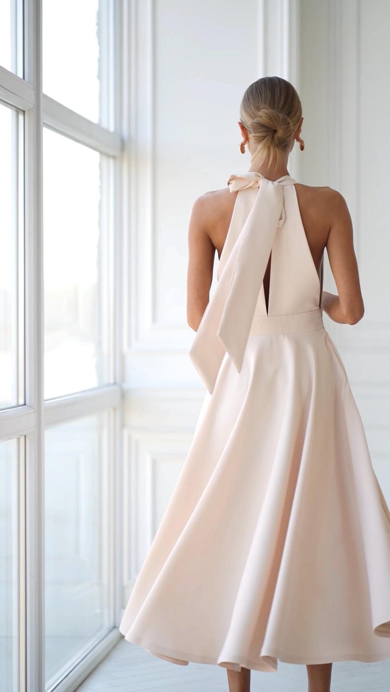 Pu&ograve; includere: Un abito a collo alto color pesca chiaro con gonna fluida e dettaglio a fiocco sulla schiena. L'abito ha una vita aderente e una gonna svasata, creando una silhouette elegante. L'abito &egrave; realizzato in un tessuto liscio e morbido.