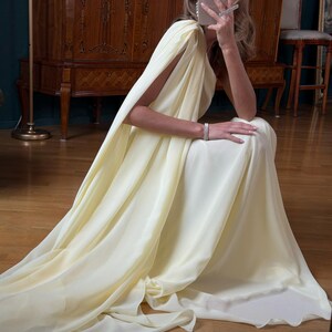 May include: A pale yellow, flowing gown with a cape-like sleeve. The dress is made of a soft, draping fabric. The person is wearing a bracelet and earrings. The background includes a wooden cabinet and a dark green wall.