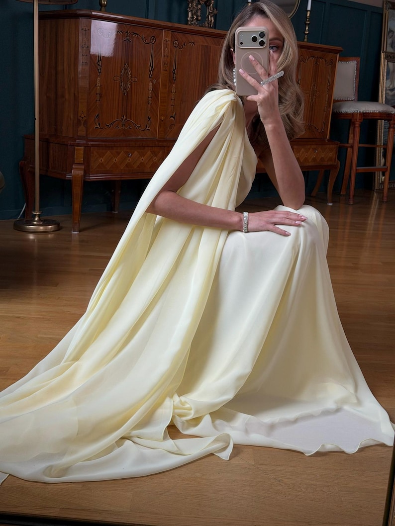 May include: A pale yellow, flowing gown with a cape-like sleeve. The dress is made of a soft, draping fabric. The person is wearing a bracelet and earrings. The background includes a wooden cabinet and a dark green wall.